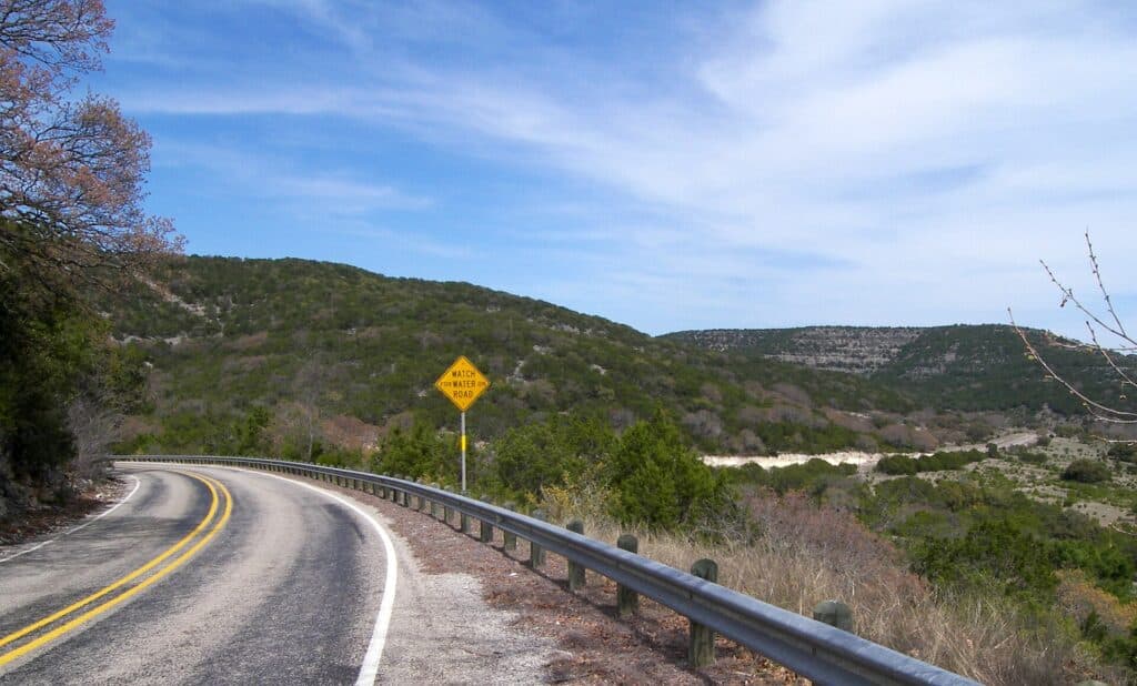A two-lane road curving through green hills in the Texas Hill Country under a blue sky